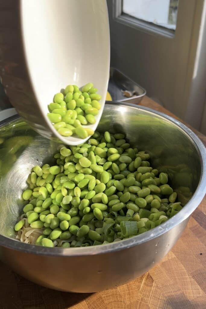 A white bowl pours bright green edamame beans and roasted corn into a large stainless steel mixing bowl on a wooden countertop, with sunlight streaming through a nearby window.