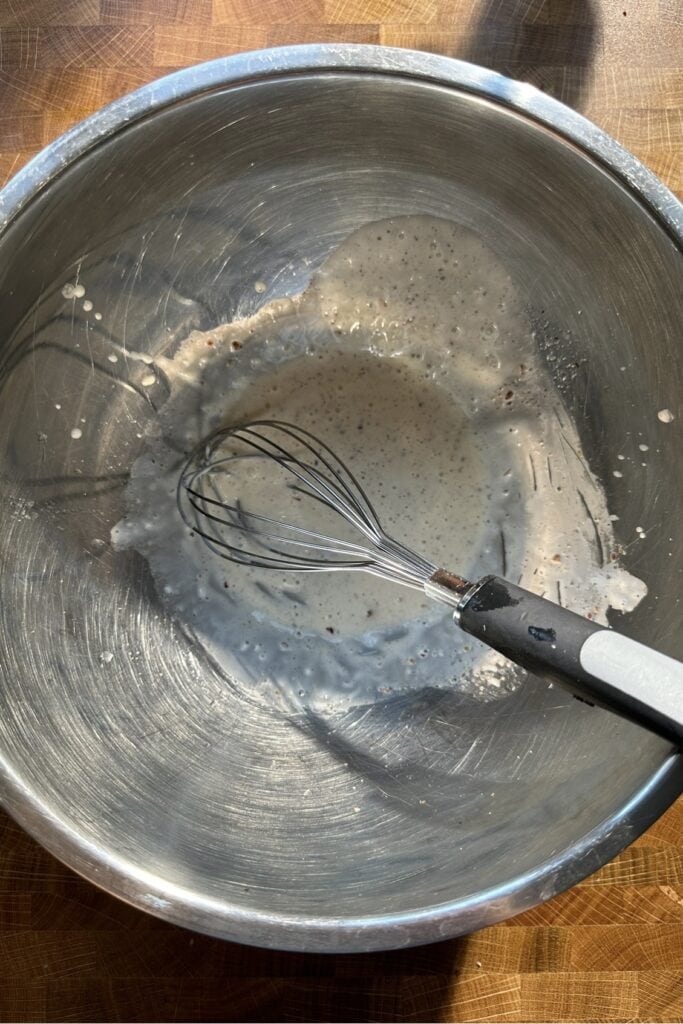 A metal mixing bowl contains a small amount of whisked roasted corn batter or sauce. A metal whisk with a black handle rests inside the bowl on a wooden surface as sunlight casts shadows across the scene.
