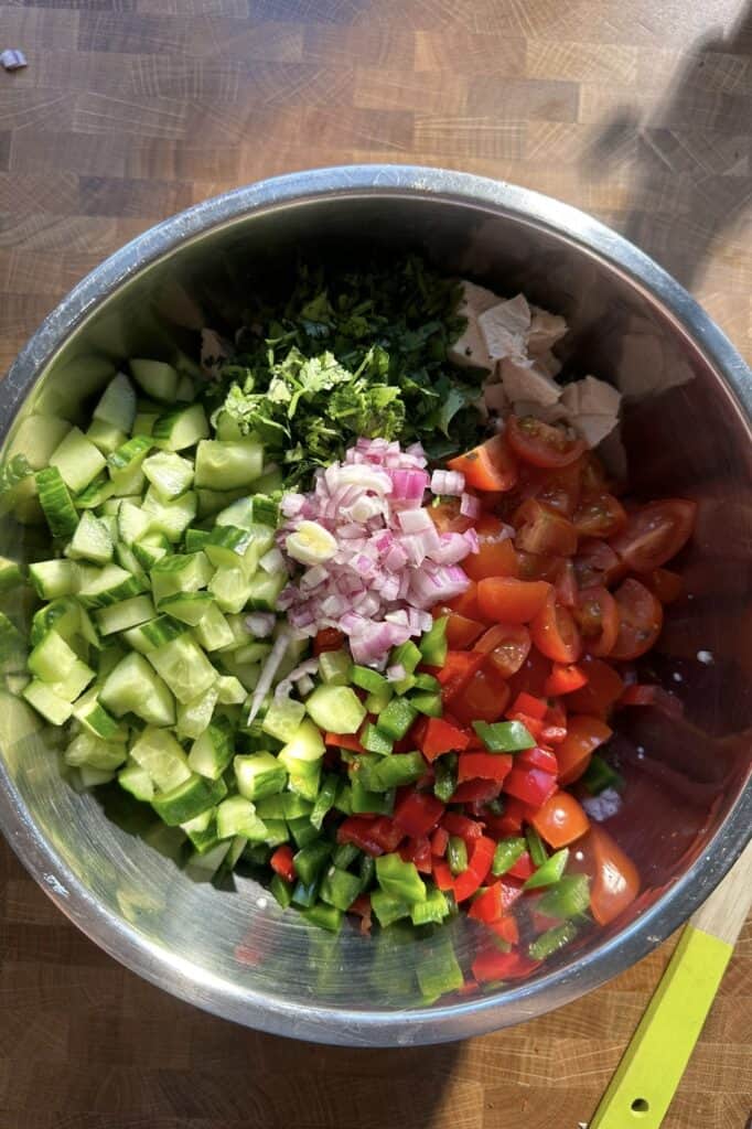 A metal bowl filled with chopped cucumber, red onion, cherry tomatoes, jalapeños, cilantro, roasted corn, and pieces of cooked chicken rests on a wooden countertop. A green knife handle is visible at the bowl’s edge.