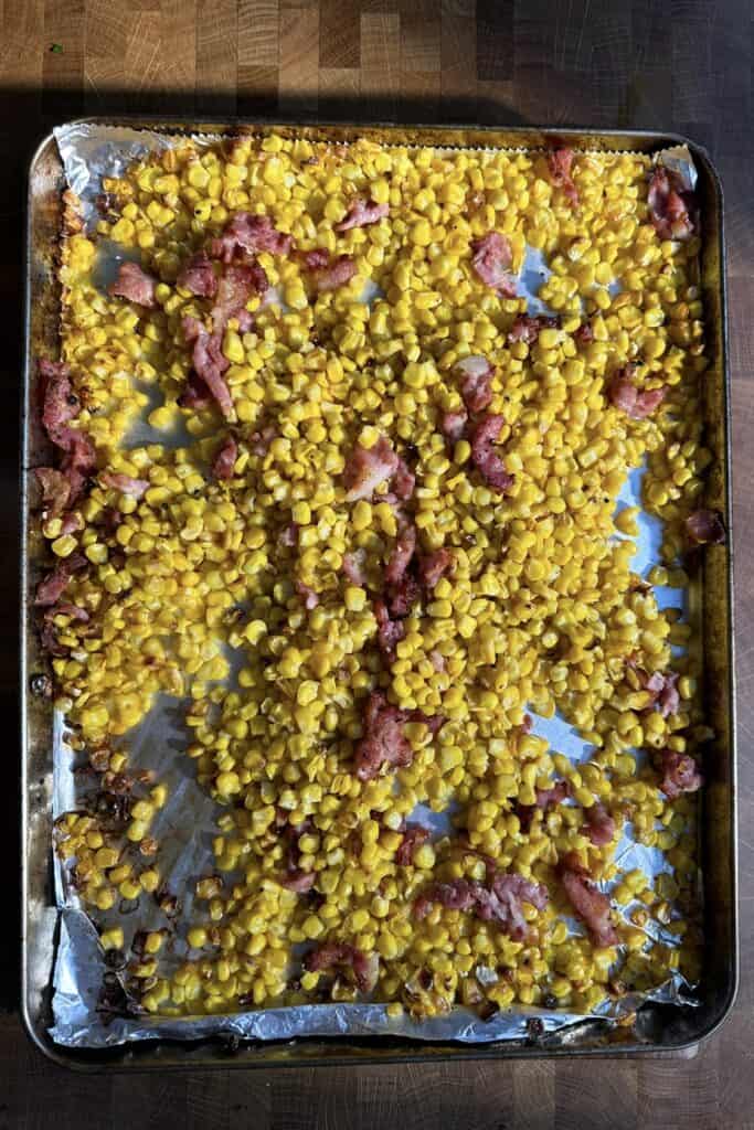 A baking sheet lined with foil holds roasted corn kernels mixed with pieces of cooked, crispy bacon on a wooden surface.