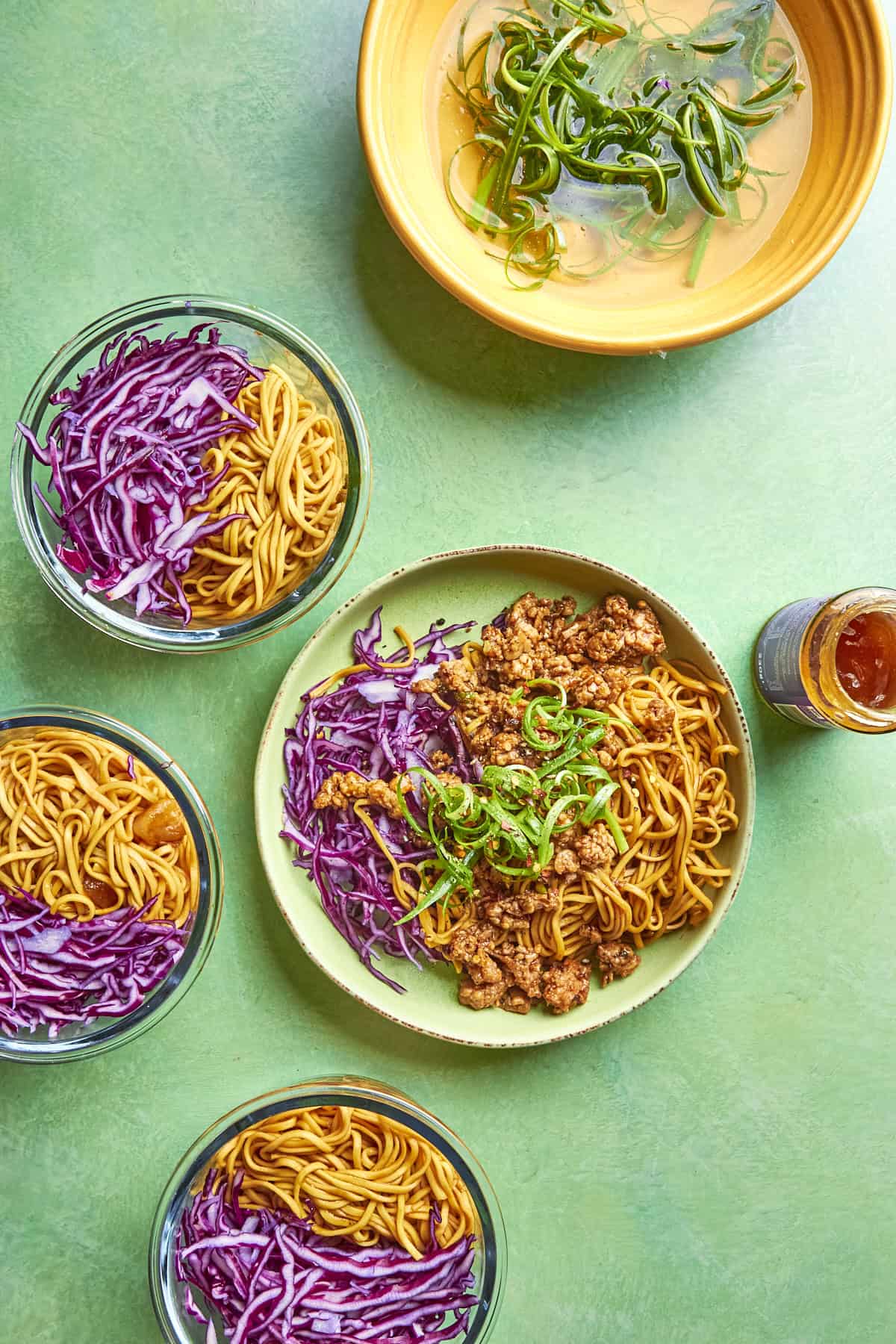 Overhead view of bowls with noodles, shredded purple cabbage, and roasted corn; a main plate with noodles, ground meat, cabbage, green onions; plus a small bowl of broth with greens—all on a green surface.