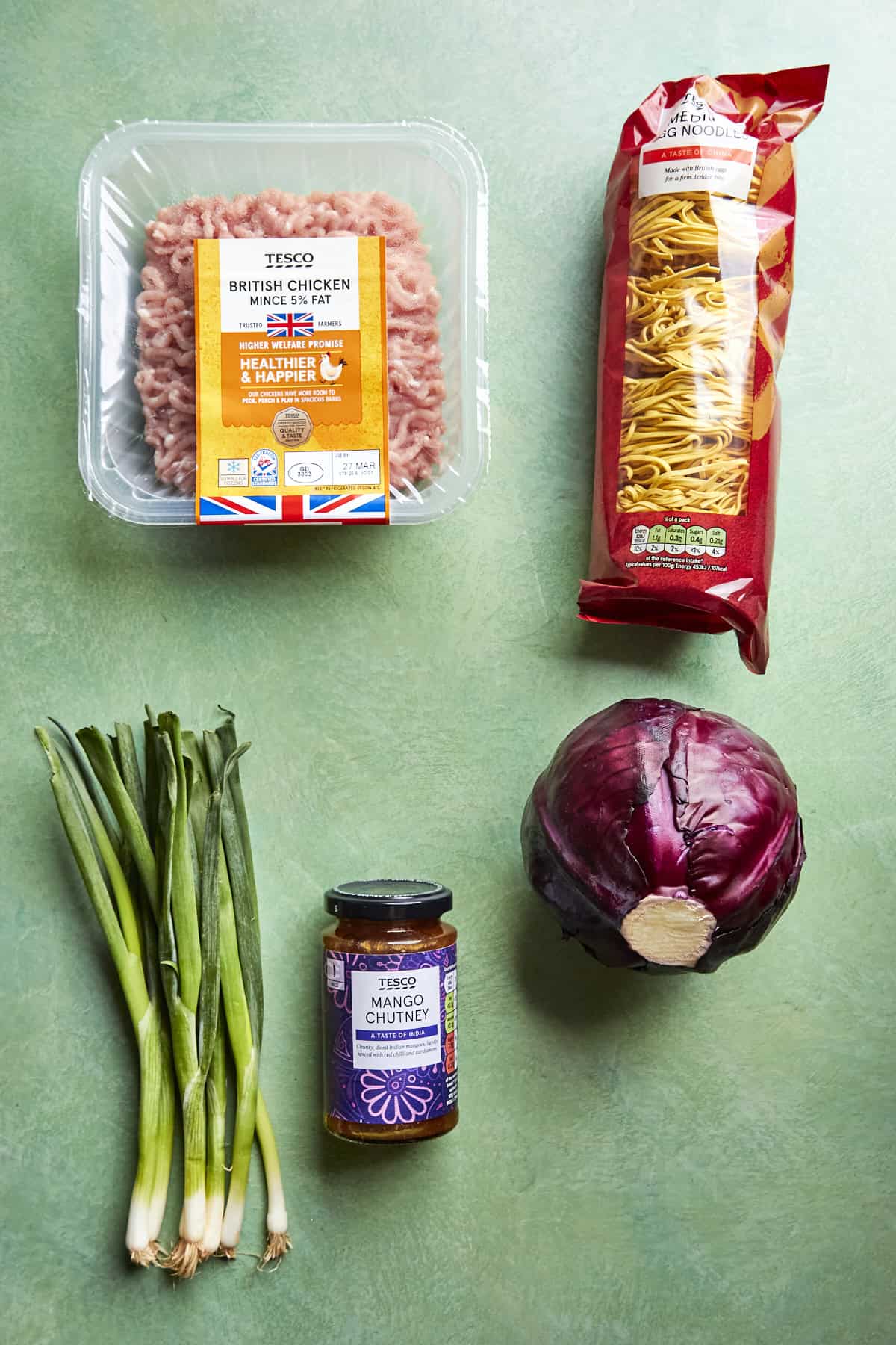 Top-down view of five ingredients on a green surface: packaged chicken mince, a pack of egg noodles, a bunch of spring onions, a jar of mango chutney, halved red cabbage, and a bowl of roasted corn.