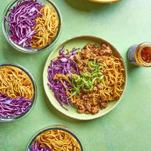 Four bowls of noodles with shredded red cabbage, and one larger bowl in the center with noodles, roasted corn, saucy ground meat, shredded cabbage, and green onions. A dish and a small jar of sauce are also visible on a green surface.