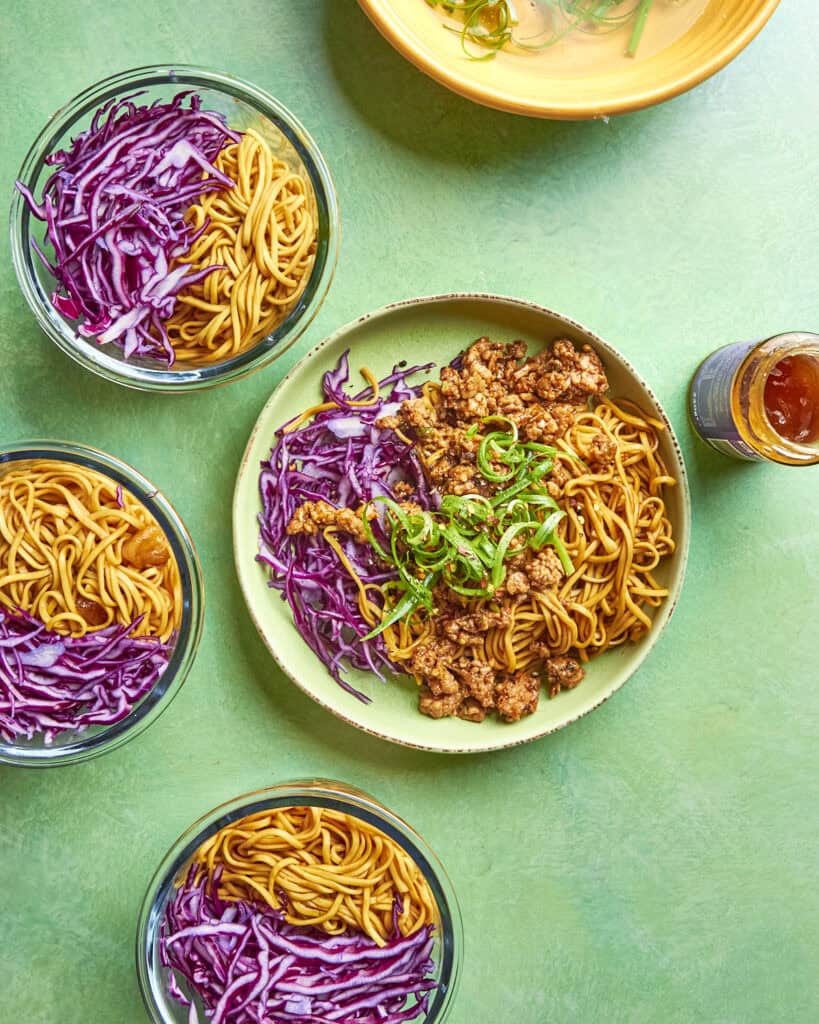 Four bowls of noodles with shredded red cabbage, and one larger bowl in the center with noodles, roasted corn, saucy ground meat, shredded cabbage, and green onions. A dish and a small jar of sauce are also visible on a green surface.
