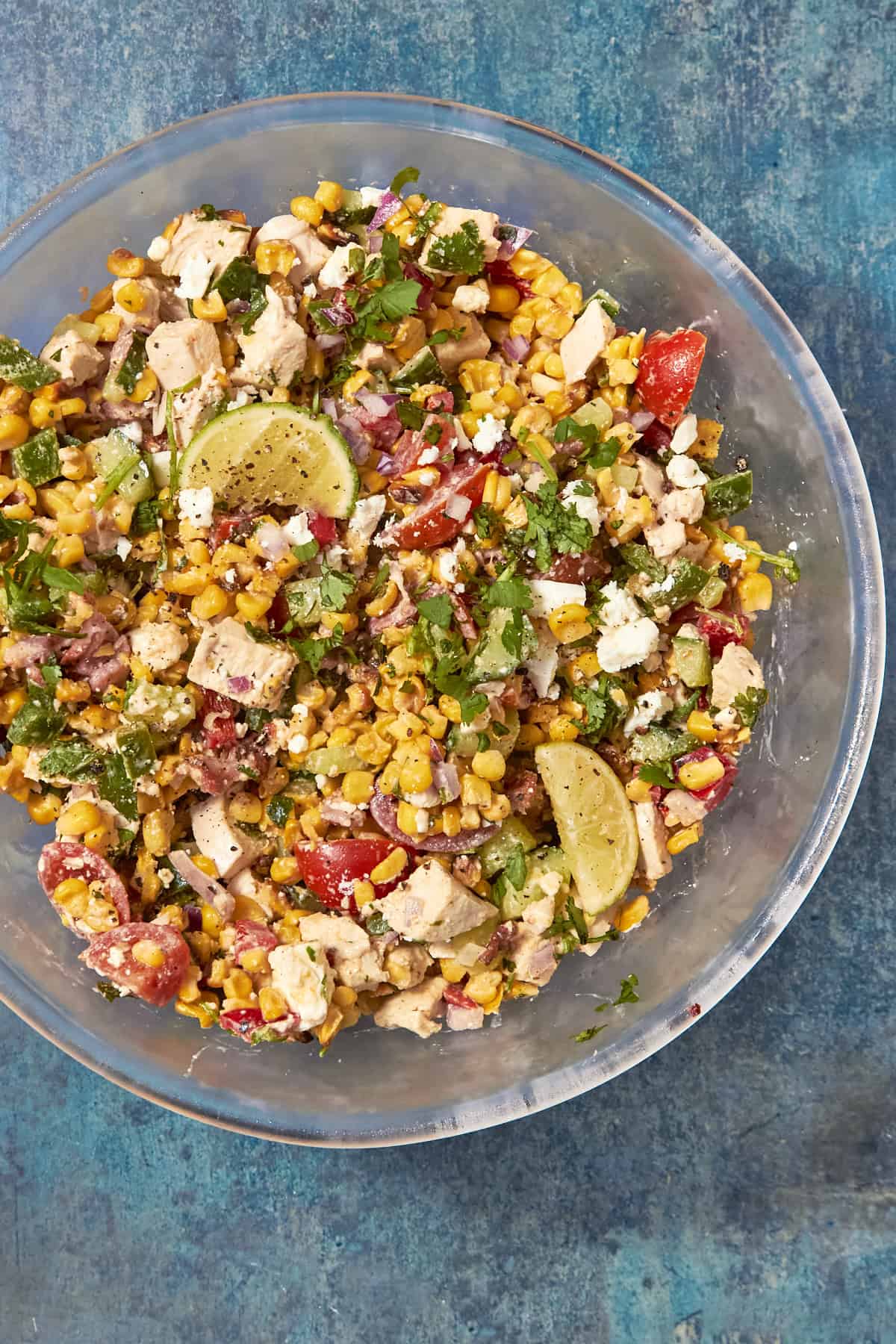 A clear bowl filled with a colorful salad of roasted corn, diced chicken, cherry tomatoes, red onion, cilantro, cheese, and lime wedges sits on a blue textured surface.