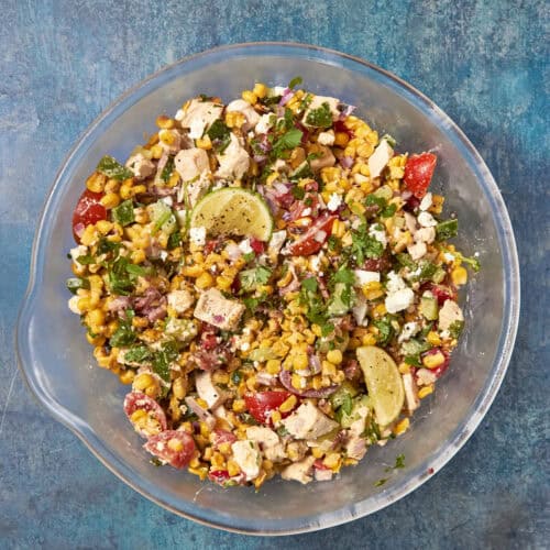 A glass bowl filled with a colorful salad containing roasted corn, chopped chicken, tomatoes, red onions, cilantro, feta cheese, and lime wedges, set on a blue textured surface.