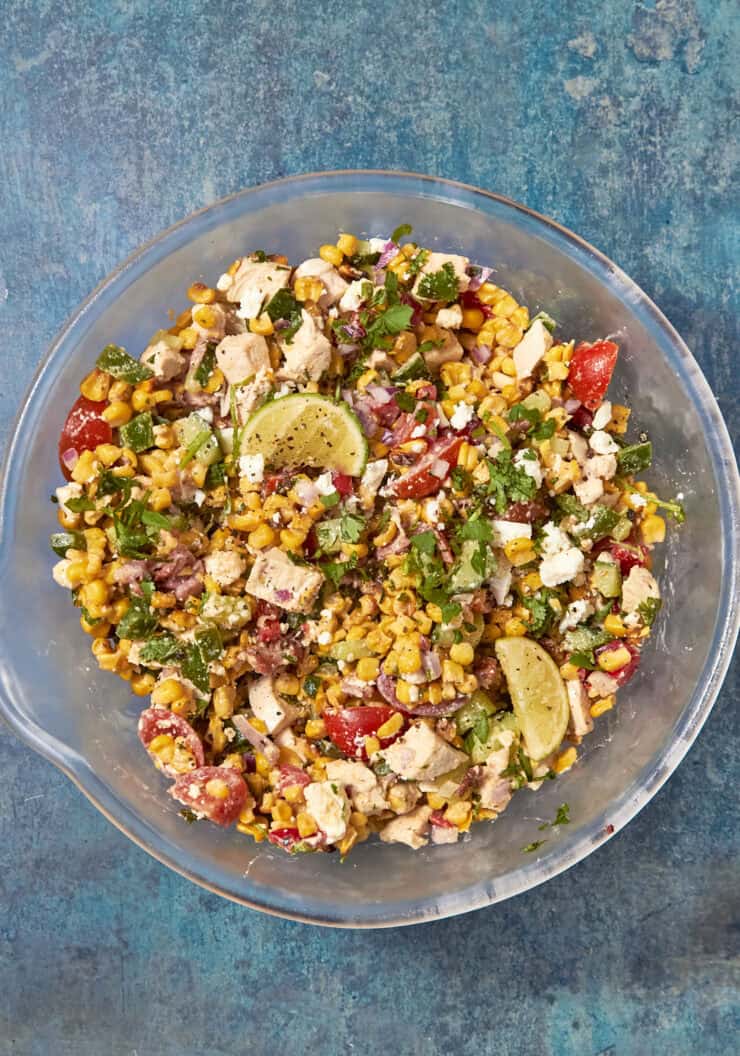 A glass bowl filled with a colorful salad containing roasted corn, chopped chicken, tomatoes, red onions, cilantro, feta cheese, and lime wedges, set on a blue textured surface.