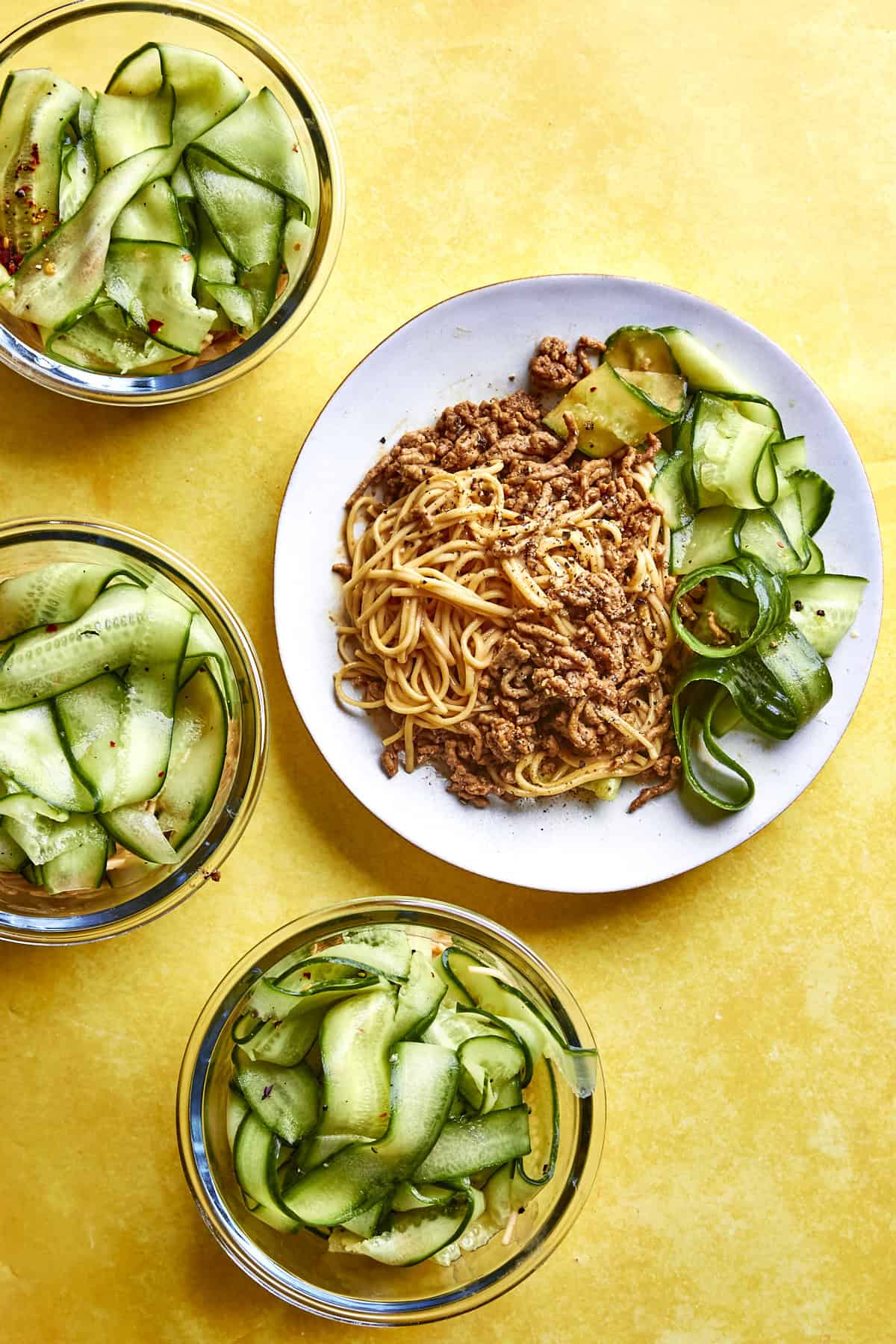 A plate of noodles topped with ground meat, roasted corn, and cucumber ribbons is surrounded by three bowls of cucumber salad on a bright yellow surface.