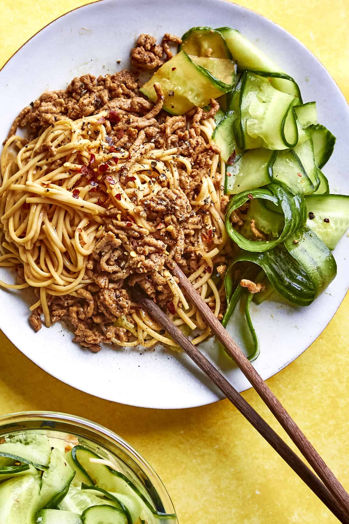 A white plate of noodles topped with seasoned ground meat and roasted corn, served with cucumber ribbons. Wooden chopsticks rest on the plate, and a small bowl of more cucumber slices is nearby on a bright yellow background.