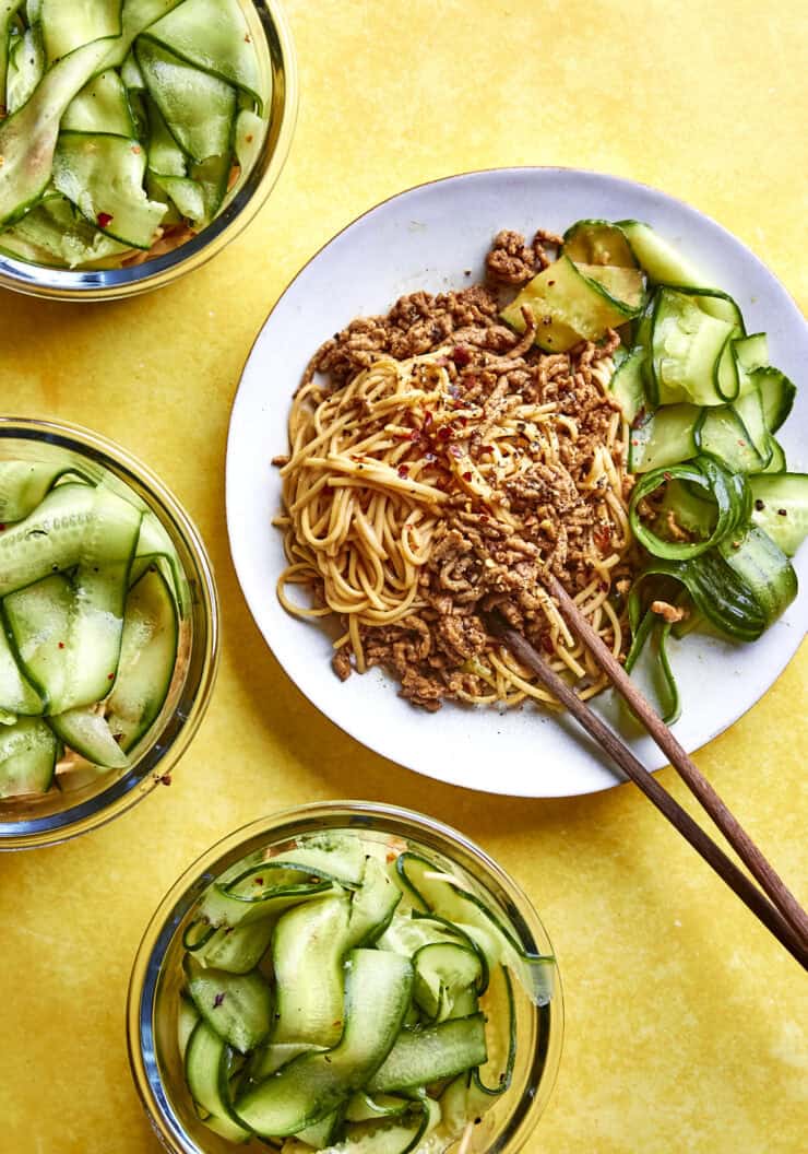 A plate of noodles topped with ground meat, roasted corn, and cucumber ribbons is served with chopsticks, surrounded by three bowls of cucumber salad on a bright yellow surface.
