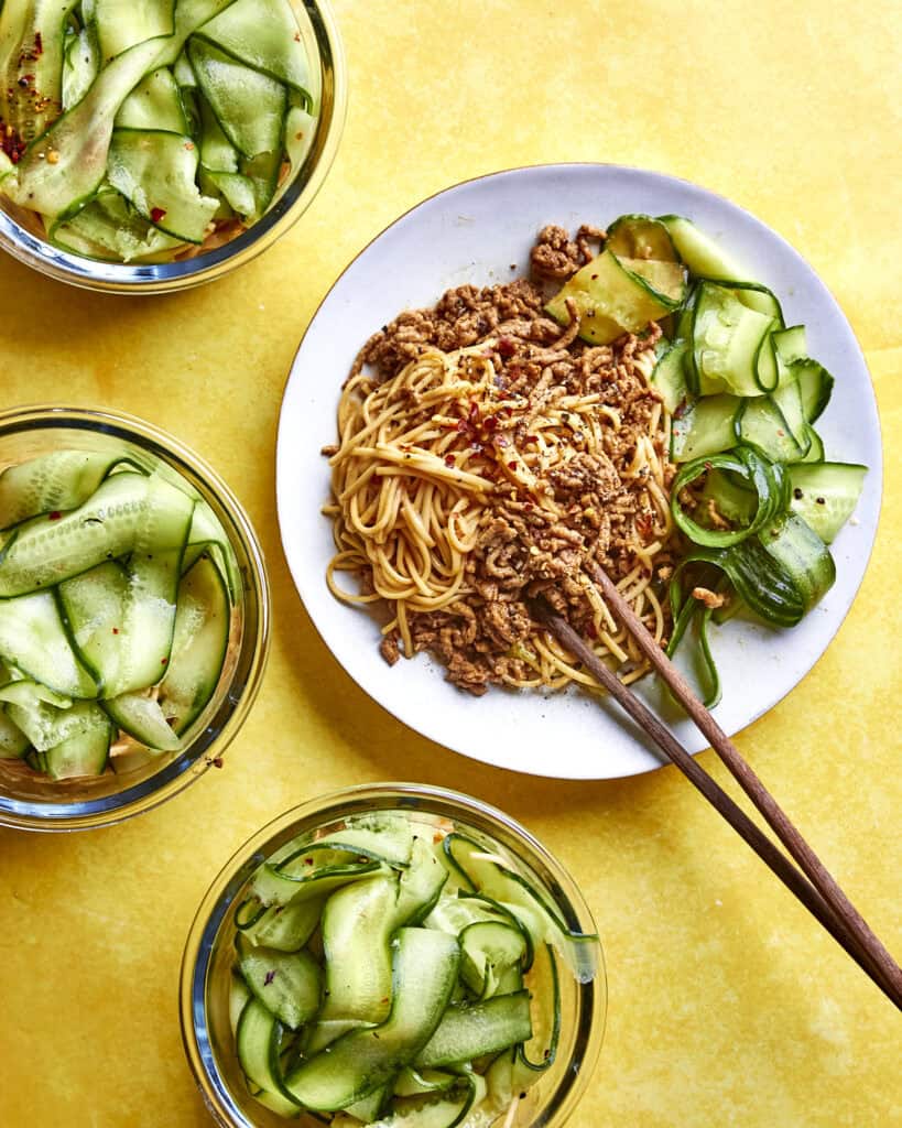 A plate of noodles topped with ground meat, roasted corn, and cucumber ribbons is served with chopsticks, surrounded by three bowls of cucumber salad on a bright yellow surface.