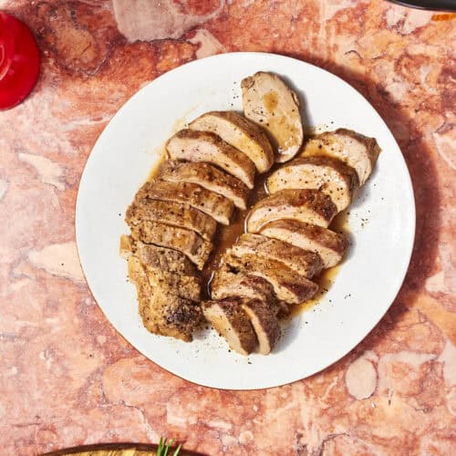 Sliced cooked pork tenderloin, from a savory Pork Loin Crock Pot Recipe, arranged on a white plate with juices. Set on a pink marble surface, it's accompanied by a wooden board with rosemary and part of a red glass nearby.