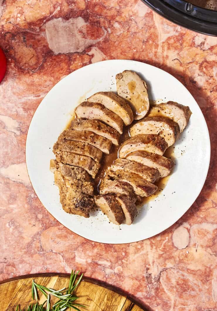 Sliced cooked pork tenderloin, from a savory Pork Loin Crock Pot Recipe, arranged on a white plate with juices. Set on a pink marble surface, it's accompanied by a wooden board with rosemary and part of a red glass nearby.