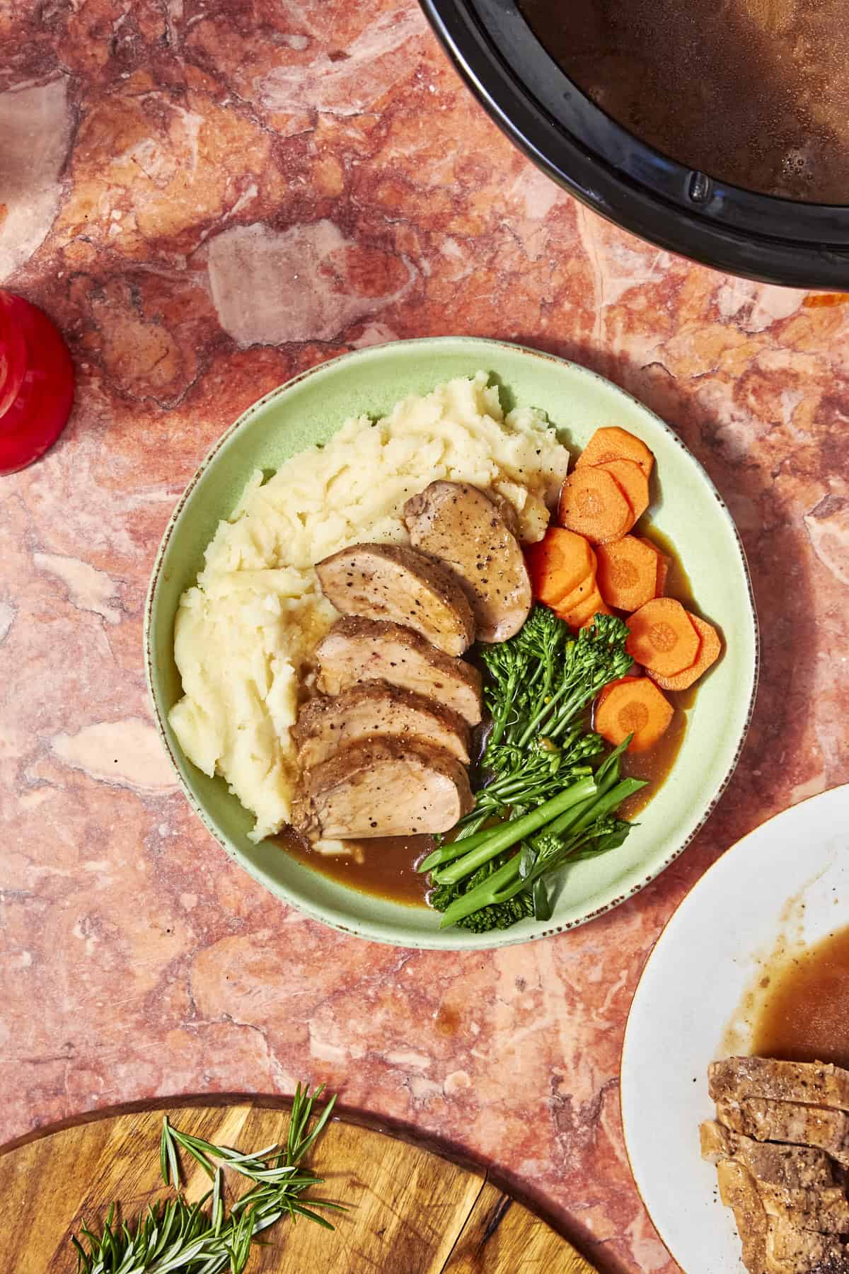 A plate of sliced roast pork with brown gravy, mashed potatoes, steamed carrots, roasted corn, and broccolini sits on a marble surface next to a slow cooker and a cutting board with rosemary.