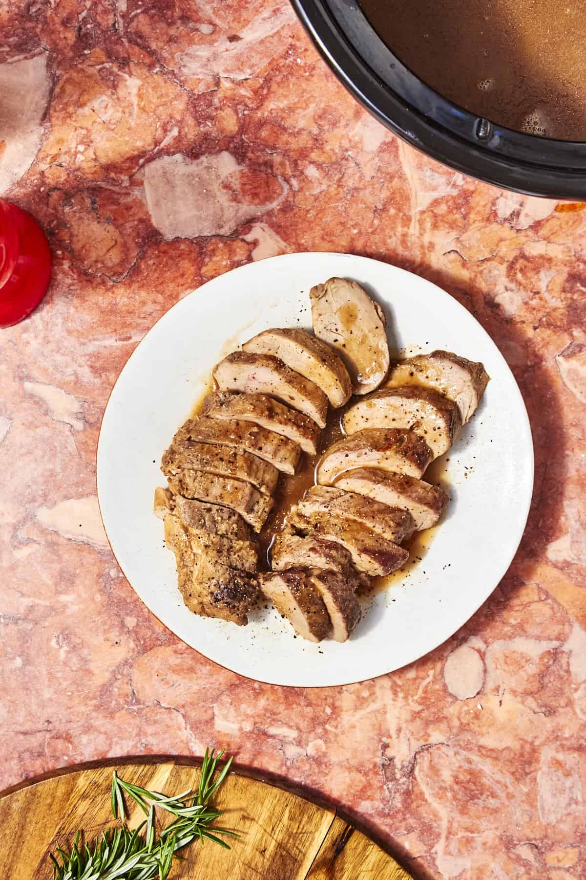Sliced cooked pork tenderloin arranged on a white plate, seasoned with pepper, on a marbled red countertop. Roasted corn and a slow cooker with sauce sit nearby, along with a wooden board topped with fresh rosemary.