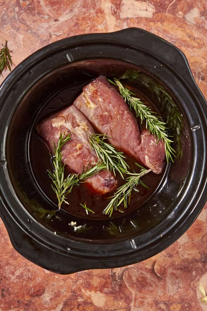 Two raw steaks marinating with sprigs of rosemary and a hint of roasted corn in a dark slow cooker, set on a reddish-brown marble surface.