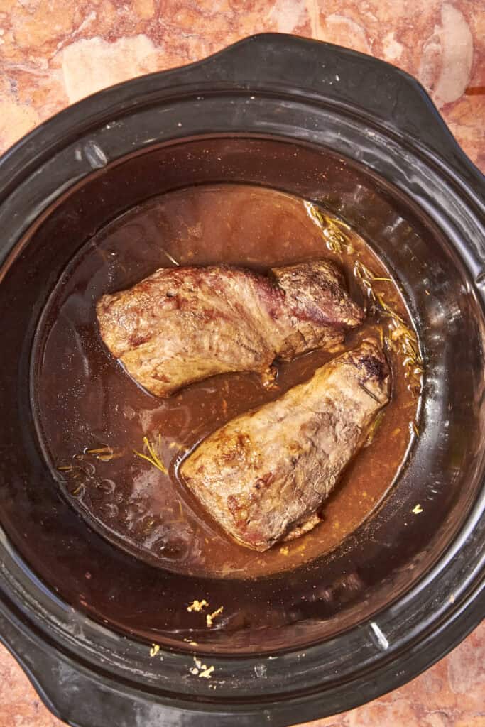 Two browned pieces of meat simmer in a rich brown sauce alongside roasted corn inside a black slow cooker, set on a marbled countertop.