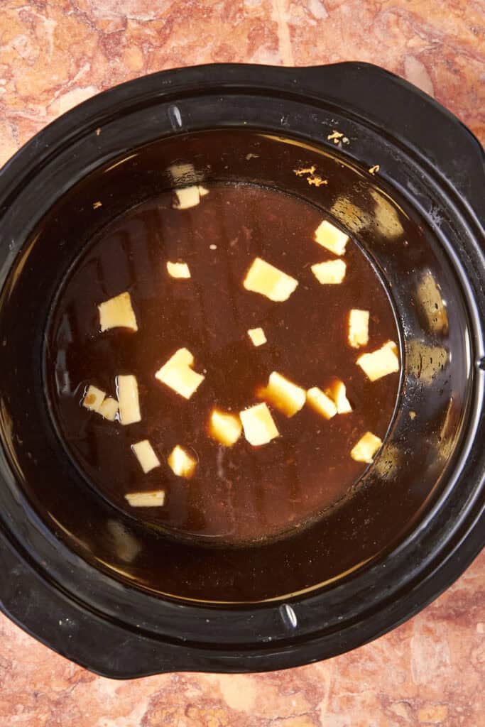 A slow cooker filled with dark brown broth, roasted corn, and several small cubes of butter floating on the surface, set on a marbled countertop.