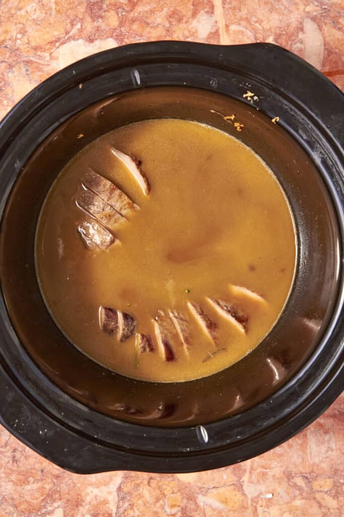 Slices of beef partially submerged in brown gravy and paired with roasted corn inside a black slow cooker, placed on a reddish-brown countertop.