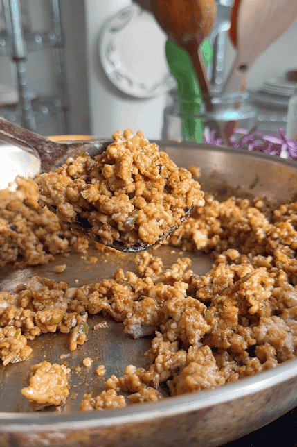 A close-up of a pan filled with seasoned cooked rice and mango sticky noodles, with a spoon lifting a portion. Wooden utensils, glass jars, and a plate are visible in the blurred background.