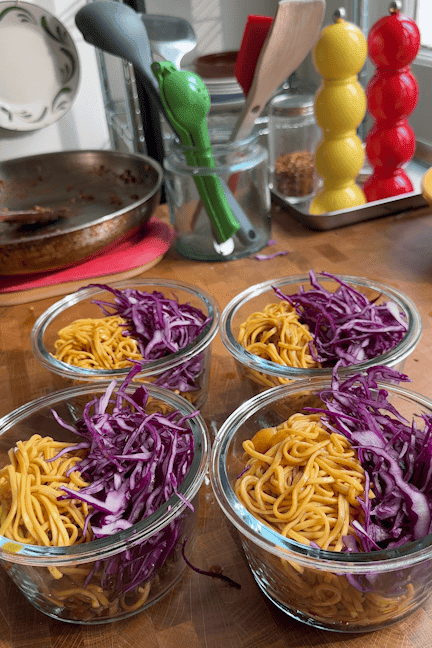Four glass bowls on a wooden counter, each filled with mango sticky noodles and shredded purple cabbage. Kitchen utensils, a pan, and colorful salt and pepper shakers are in the background.