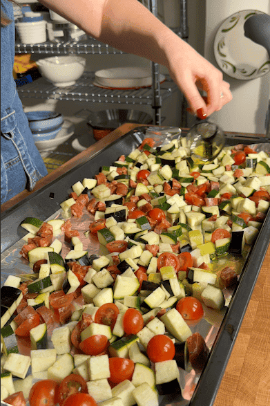 A person drizzles olive oil over a baking tray filled with chopped zucchini, eggplant, cherry tomatoes, sausage pieces, and roasted corn, preparing vegetables for roasting in a kitchen.