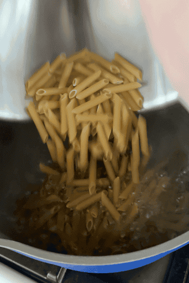 Uncooked penne pasta being poured from a metal bowl into a pot of boiling water on a stovetop, ready to be served with roasted corn for a delicious meal.