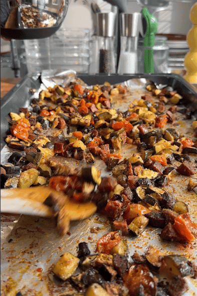 A close-up of a foil-lined baking tray filled with roasted diced vegetables, including tomatoes, zucchini, and roasted corn, being stirred with a wooden spoon in a brightly lit kitchen.