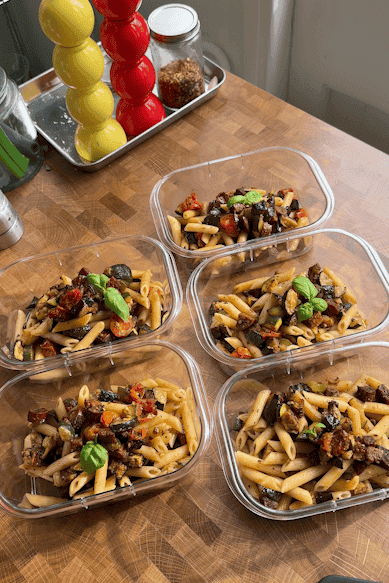 Five meal prep containers filled with penne pasta, roasted vegetables, roasted corn, and basil leaves are arranged on a wooden countertop. Colorful spice shakers and a jar of chili flakes are in the background.