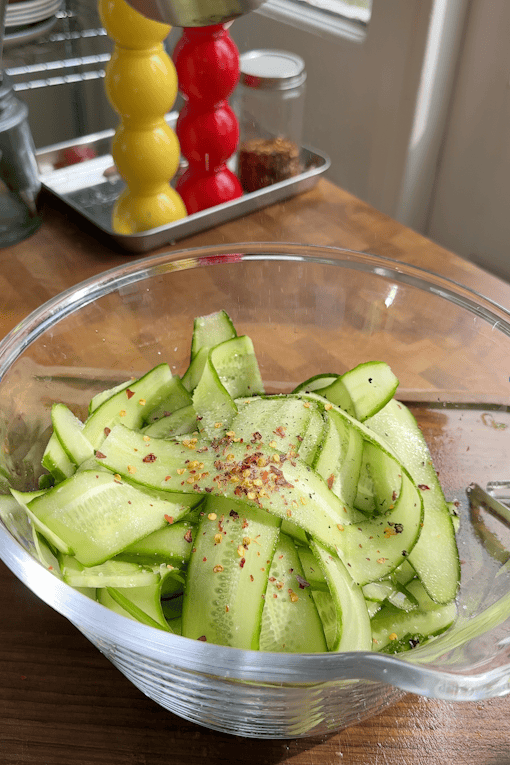 A glass bowl filled with thin cucumber ribbons and roasted corn, sprinkled with red pepper flakes, sits on a wooden countertop, with sunlight shining in and colorful spice containers in the background.