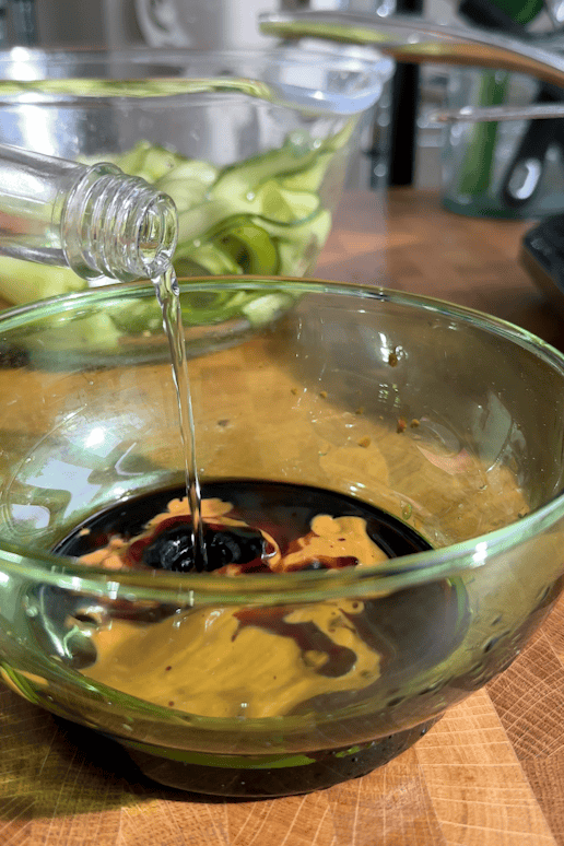 A clear liquid is being poured from a bottle into a glass bowl containing soy sauce, mustard, and roasted corn. In the background, another bowl holds cucumber slices. Both bowls are on a wooden surface.