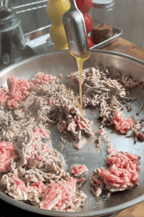 Ground meat browning in a stainless steel pan, with oil being poured over it from a metal spout. In the background, there are jars of spices, a pepper grinder, a salt grinder, and a bowl of roasted corn.