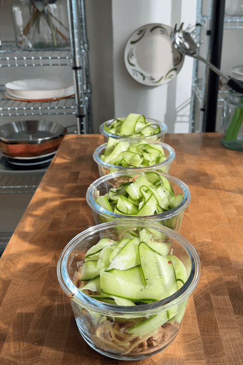 Four glass containers filled with food, each topped with thinly sliced zucchini ribbons and roasted corn, are lined up on a wooden countertop in a sunlit kitchen with shelves and dishes in the background.
