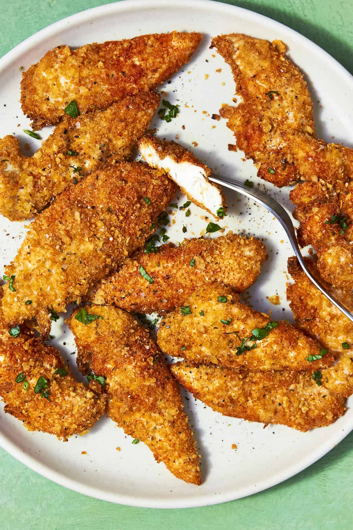 A plate of golden-brown, crispy Shake and Bake Chicken tenders garnished with chopped herbs. One tender is cut open with a fork, revealing juicy white meat inside. The plate sits on a light green surface.