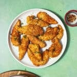 A white plate with crispy, golden-brown Shake and Bake Chicken tenders garnished with chopped herbs, set on a green table beside bowls of cornflakes, red pepper flakes, and green herbs.