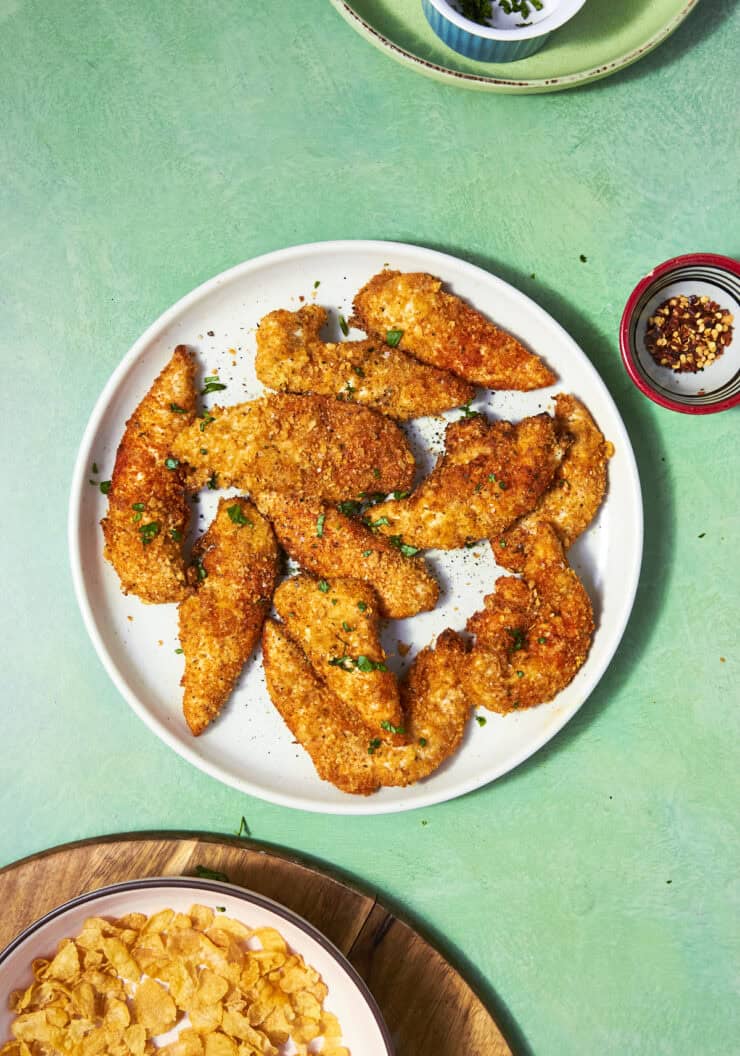 A white plate with crispy, golden-brown Shake and Bake Chicken tenders garnished with chopped herbs, set on a green table beside bowls of cornflakes, red pepper flakes, and green herbs.