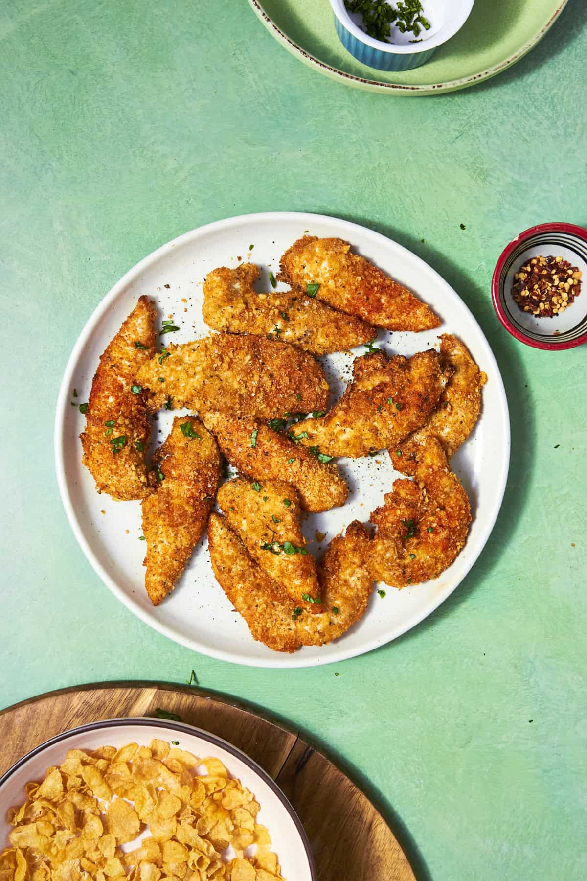 A white plate with crispy, golden-brown Shake and Bake Chicken tenders garnished with chopped herbs, set on a green table beside bowls of cornflakes, red pepper flakes, and green herbs.