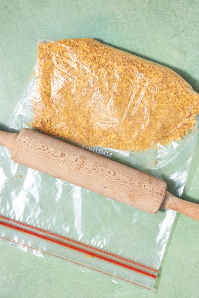 A rolling pin crushes cornflakes inside a clear plastic zip-top bag on a green surface, perfect for making crunchy Shake and Bake Chicken.