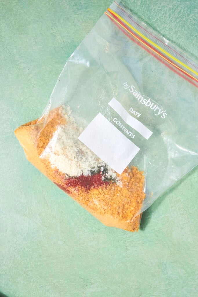 A clear ziplock bag labeled Sainsburys containing separate piles of different powdered spices or Shake and Bake Chicken seasonings, placed on a green textured surface.