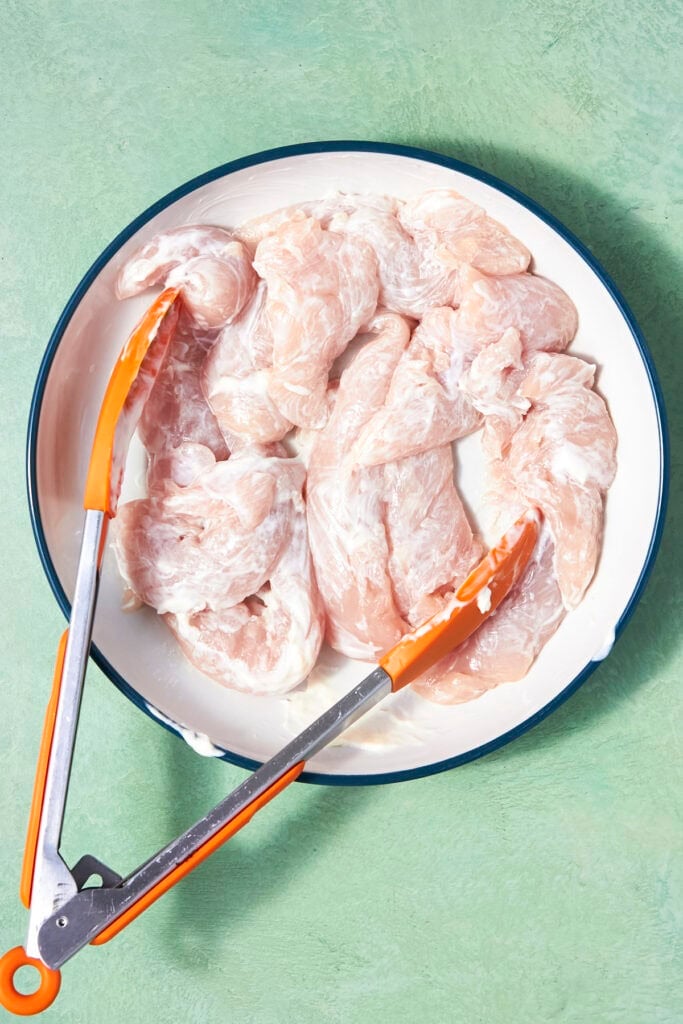 A white plate with raw Shake and Bake Chicken pieces coated in a white marinade, placed on a green surface. Orange-handled tongs rest on the edge of the plate.