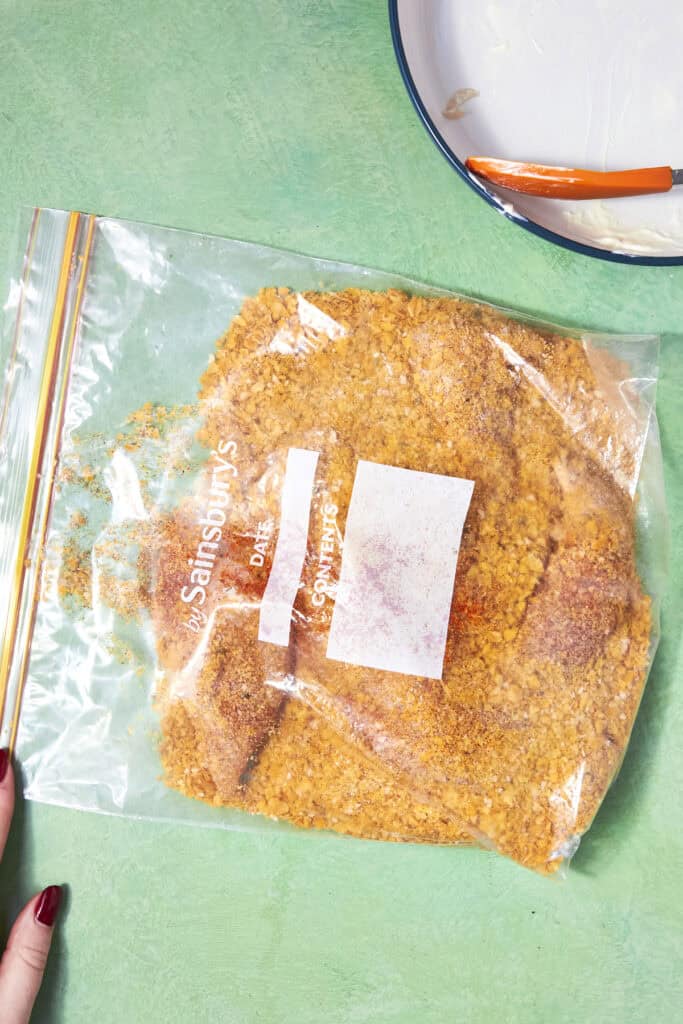 A ziplock bag filled with seasoned Shake and Bake Chicken breading mix and pieces of raw chicken rests on a green surface. A white bowl with a red spatula and some residue is partially visible in the top right corner, with a hand at the bottom left.