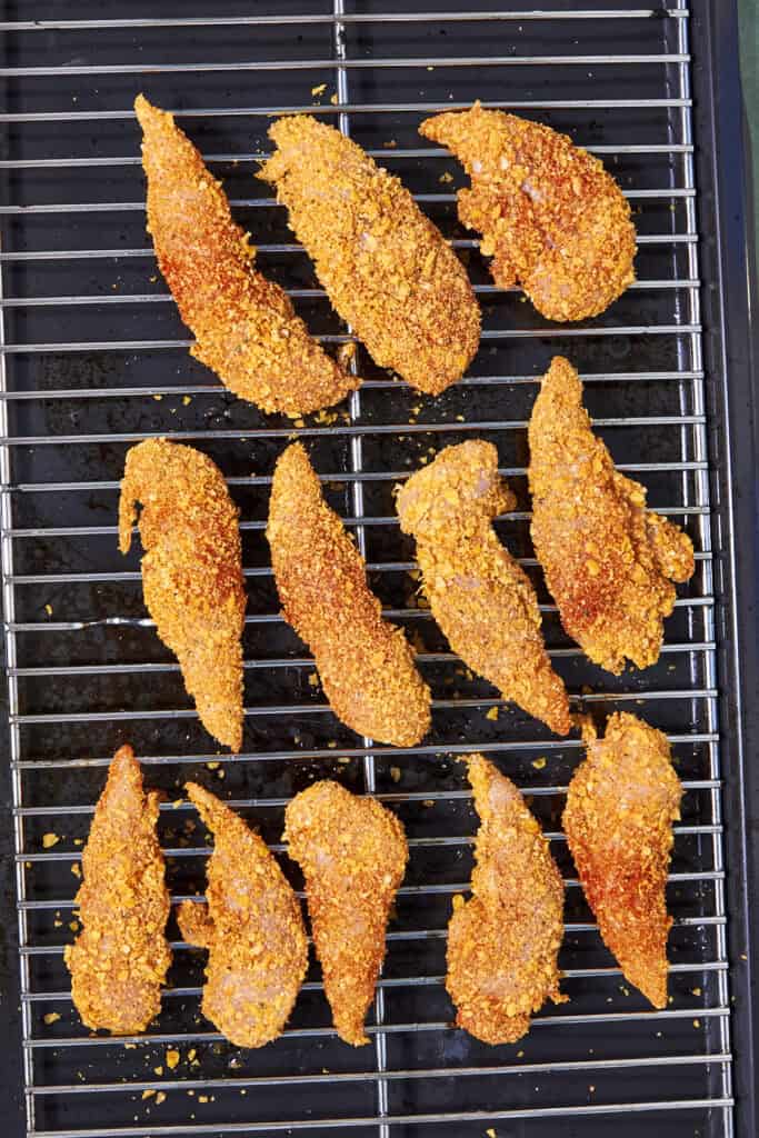 Twelve Shake and Bake Chicken tenders are arranged on a wire rack over a baking tray, ready to be cooked. The tenders are coated in a golden, crispy crust.