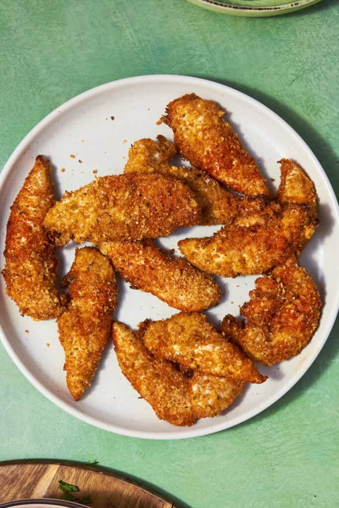 A white plate filled with crispy, golden-brown Shake and Bake Chicken tenders sits on a green surface.