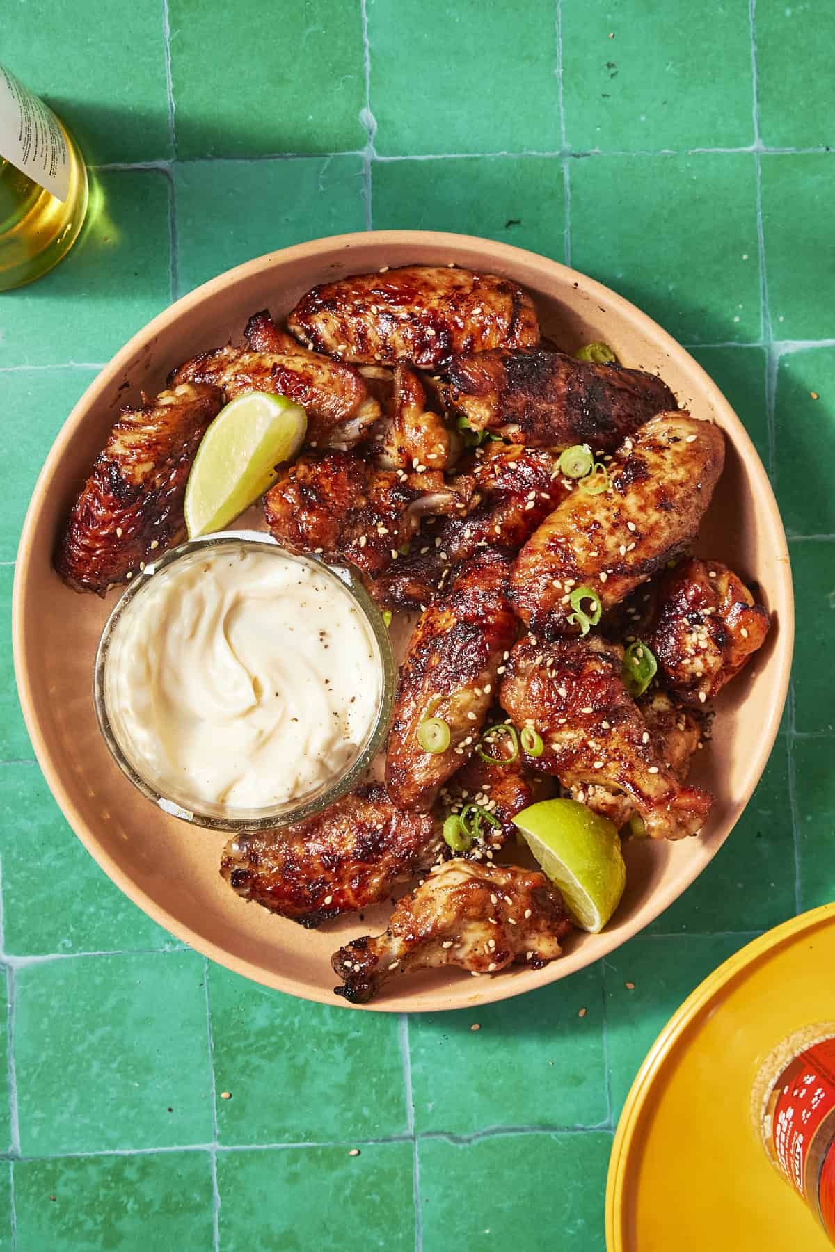 A bowl of soy chicken wings garnished with green onions and lime wedges, served with a side of creamy dipping sauce on a bright green tiled surface.