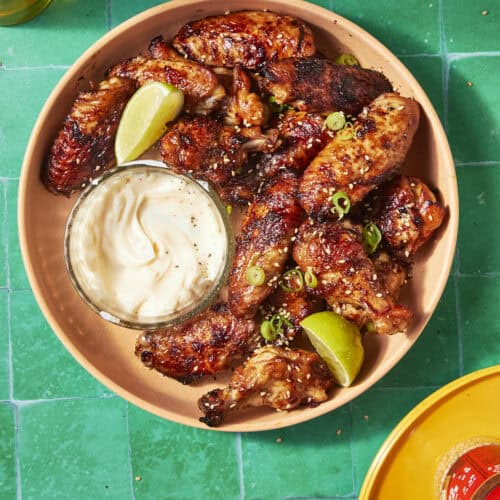 A plate of soy chicken wings garnished with sliced green onions and lime wedges, served with a bowl of creamy dipping sauce, sits on a bright green tiled surface.