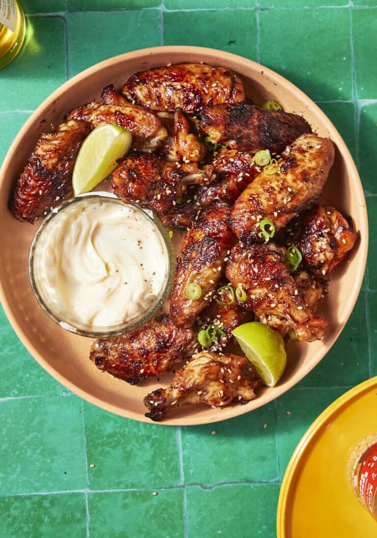 A plate of soy chicken wings garnished with sliced green onions and lime wedges, served with a bowl of creamy dipping sauce, sits on a bright green tiled surface.