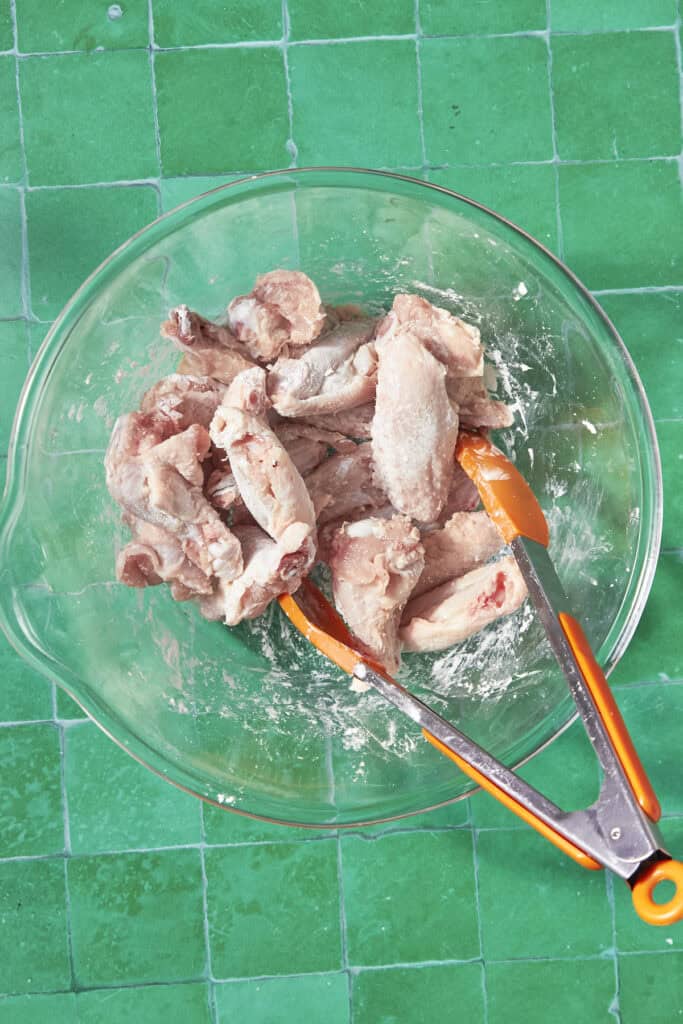 A glass bowl containing raw, floured soy chicken wings sits on a green tiled surface. Orange-handled tongs rest in the bowl, touching the wings.
