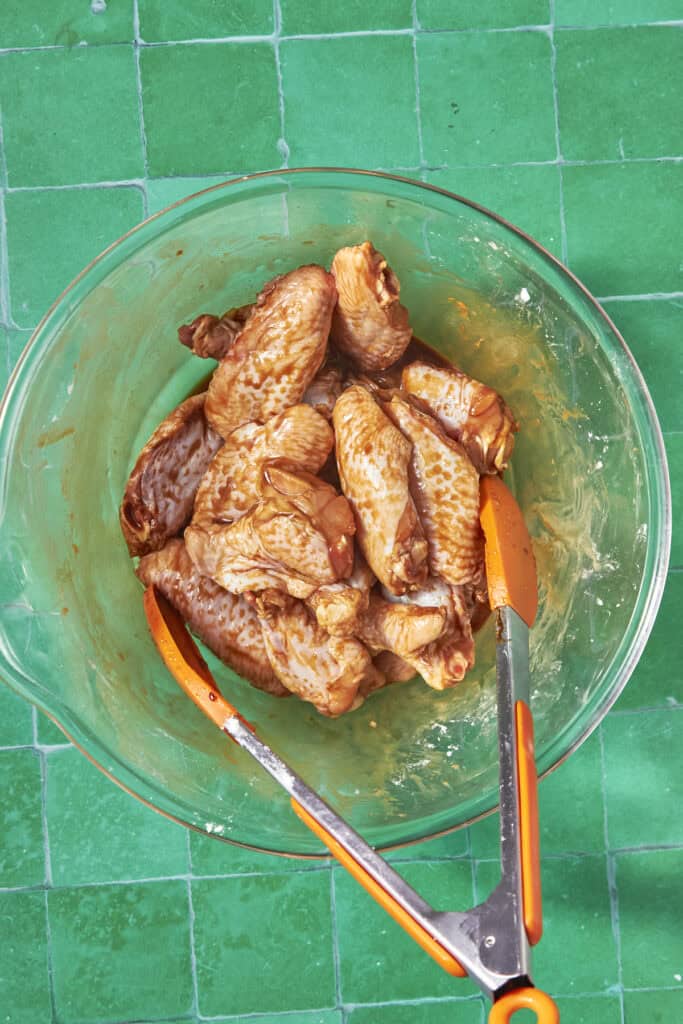 A glass bowl filled with marinated soy chicken wings sits on a green tiled surface, with a pair of orange-handled tongs resting inside the bowl.