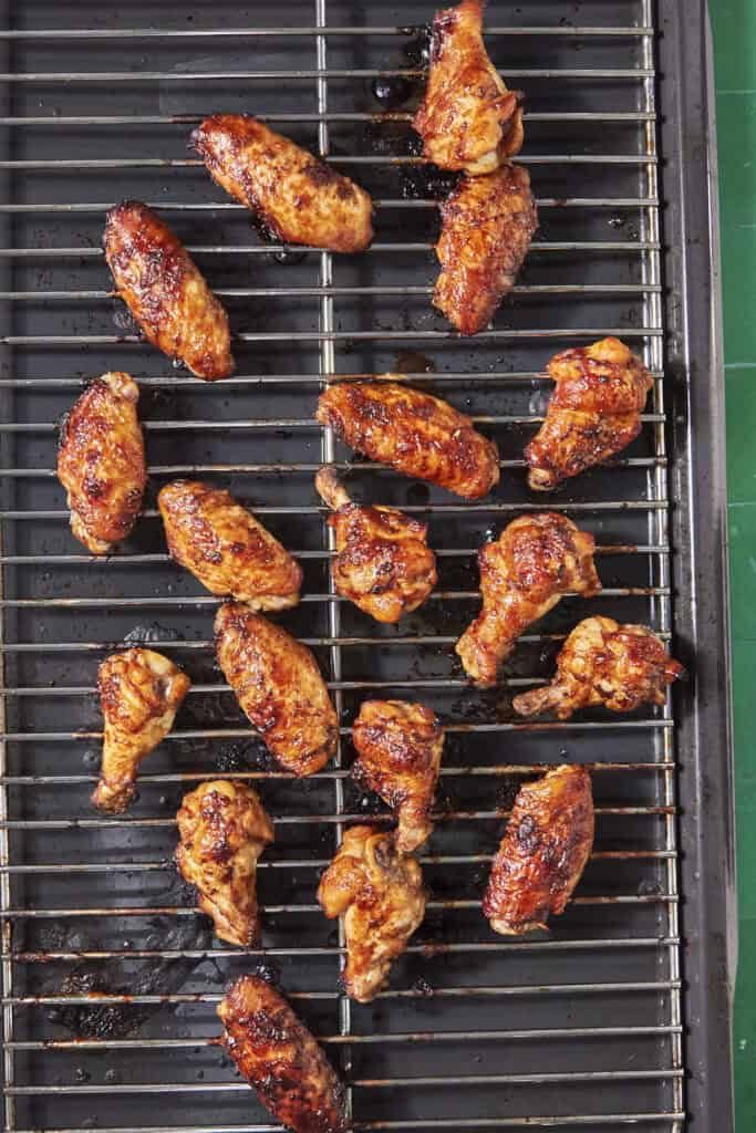 Soy chicken wings are coated in a shiny, caramelized sauce and arranged on a metal grill rack, with visible grill marks and some charred spots. The grill sits over a drip tray collecting savory juices.