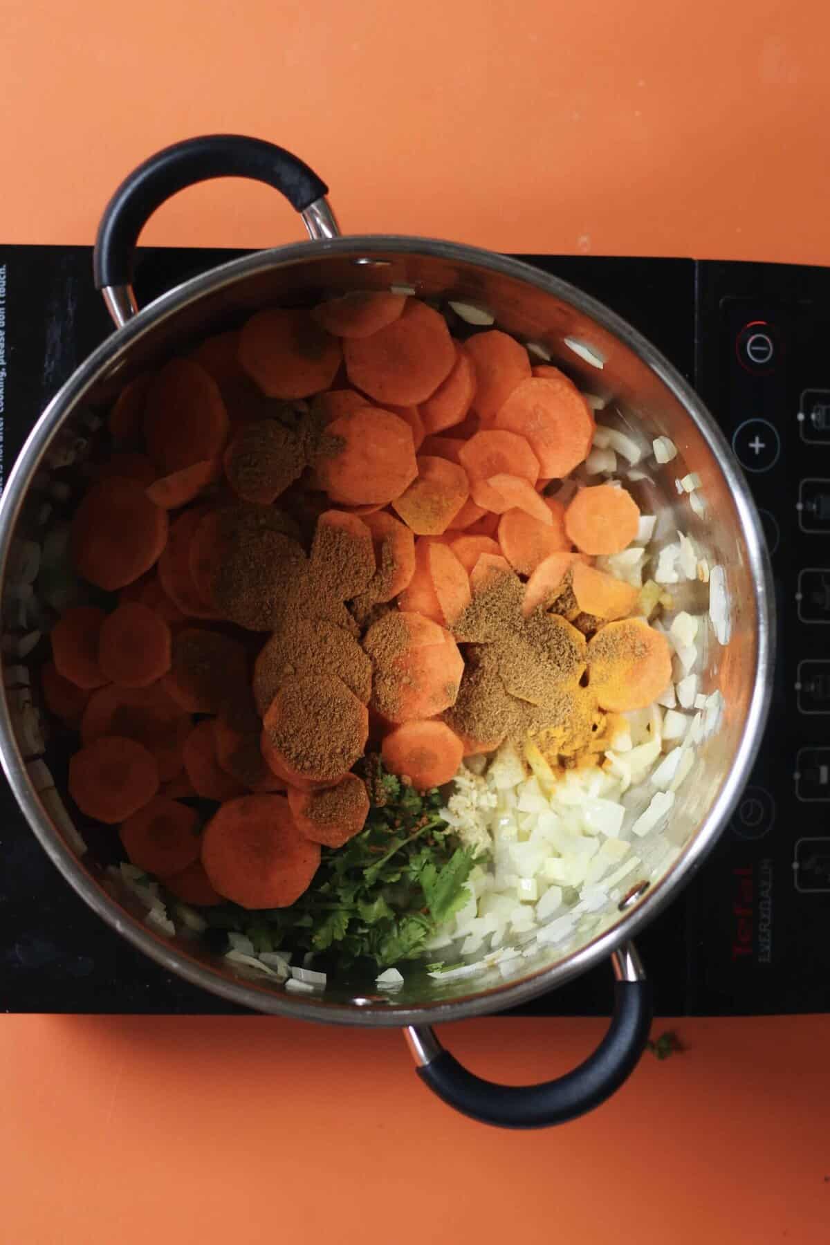 Carrots, spices, onions, lentils and coriander in large saucepan on a stove on an orange background.